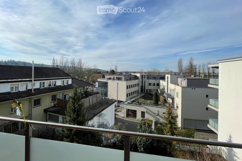 Balcony overlooking a residential area with multiple buildings, trees, and a clear sky.