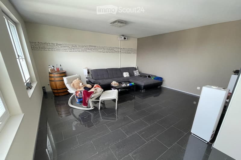 Modern living room with dark tiled flooring, large grey sectional sofa, coffee table, white rocking chair with teddy bear, barrel side table, and decorative wall with movie titles.
