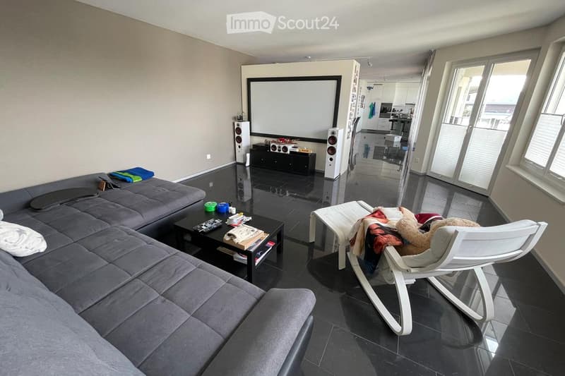 Modern living room with a grey sectional sofa, coffee table, white chair, and a large screen. The room features dark flooring and large windows with blinds.