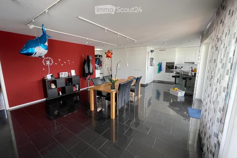 Dining area with a wooden dining table and chairs, a black cabinet with picture frames, balloons, and a kitchen area with white cabinets and stools.