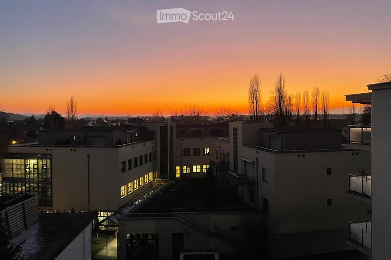A multi-story building complex with illuminated windows, surrounded by trees and a cityscape at sunset.