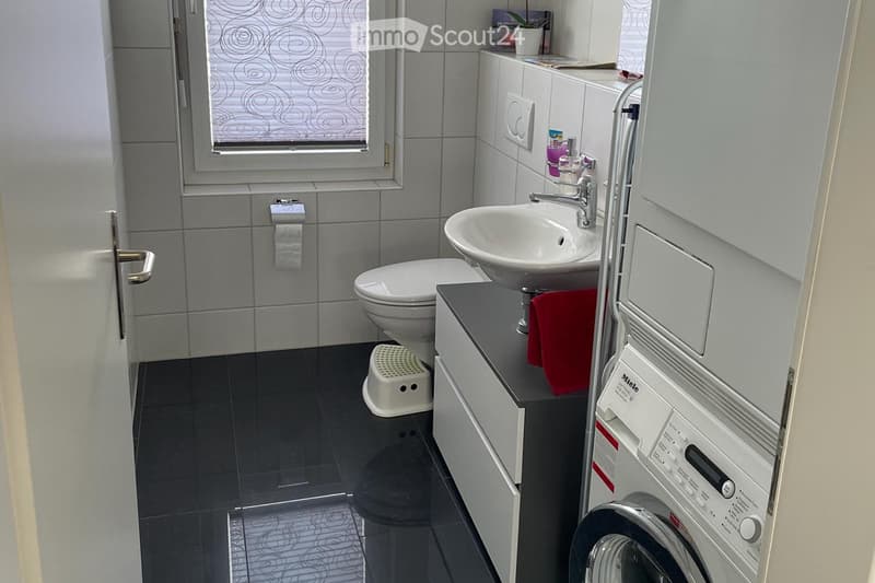 Modern bathroom with a toilet, sink, faucet, cabinet, towel, washing machine, and a small stool. The room features dark tiled flooring and grey tiled walls.