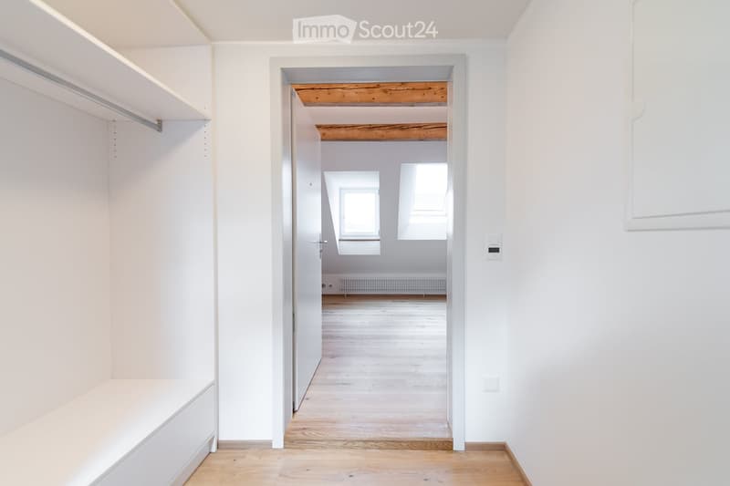 An empty room with white walls and wooden flooring, featuring built-in shelves on one side and a doorway leading to another room with exposed wooden beams and windows.