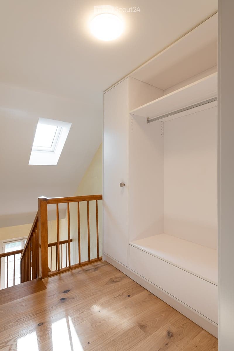 Bright hallway with wooden flooring, white built-in closet with shelves and drawers, wooden staircase railing, and a skylight window providing natural light.