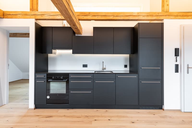 Modern kitchen with dark grey cabinets, stainless steel sink, and built-in oven. Exposed wooden beams on the ceiling and light wooden flooring.
