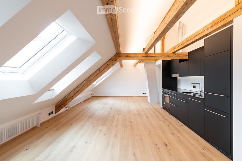 Bright, open attic space with wooden beams and skylights, featuring a modern kitchen area with dark cabinets and a sink.