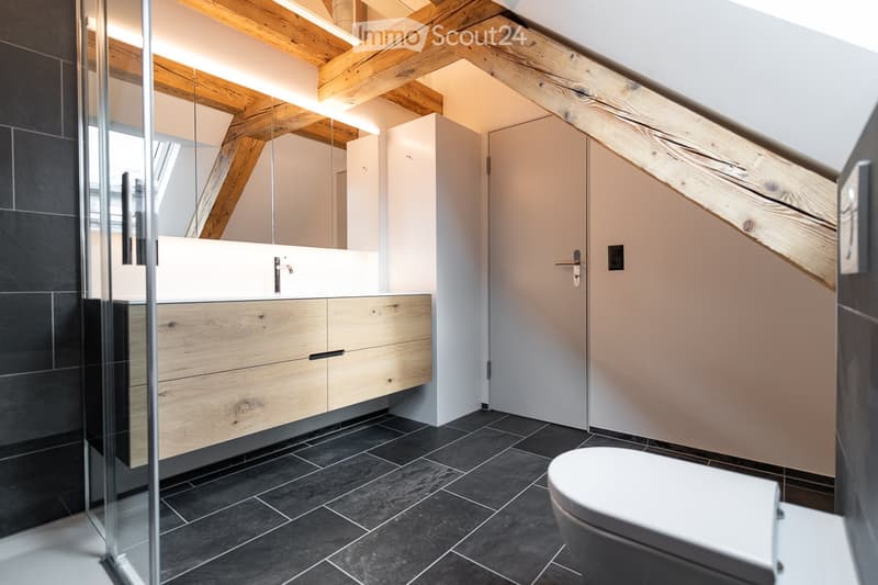 Modern bathroom with dark tiled floor, wooden cabinetry, white sink, and a toilet. Exposed wooden beams on the ceiling and a glass shower enclosure.