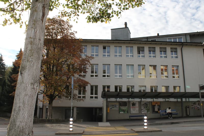 A multi-story building with a light-colored facade and numerous windows, featuring a small bus stop shelter with benches and posters on the wall.