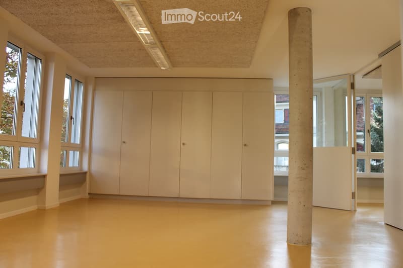 Empty room with light-colored walls, large windows, built-in cabinetry, and a central concrete pillar. The floor is a smooth, light-colored material, and the space is well-lit with natural light from the windows.