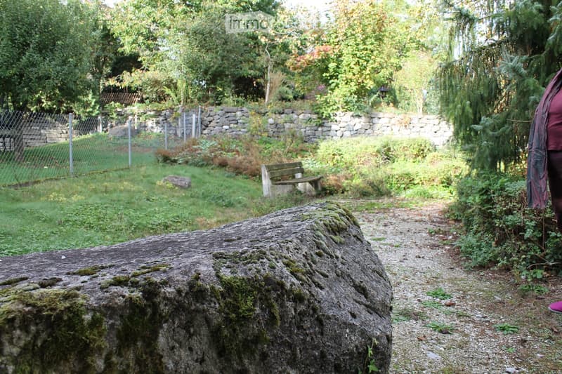 A garden area with a large moss-covered stone, a wooden bench, a stone wall, and a fenced area with trees and greenery.