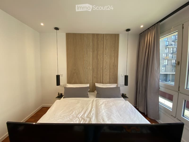 Modern bedroom with a large bed, wooden headboard, grey pillows, nightstands, and pendant lights. Large window with curtains offers a view of the city.