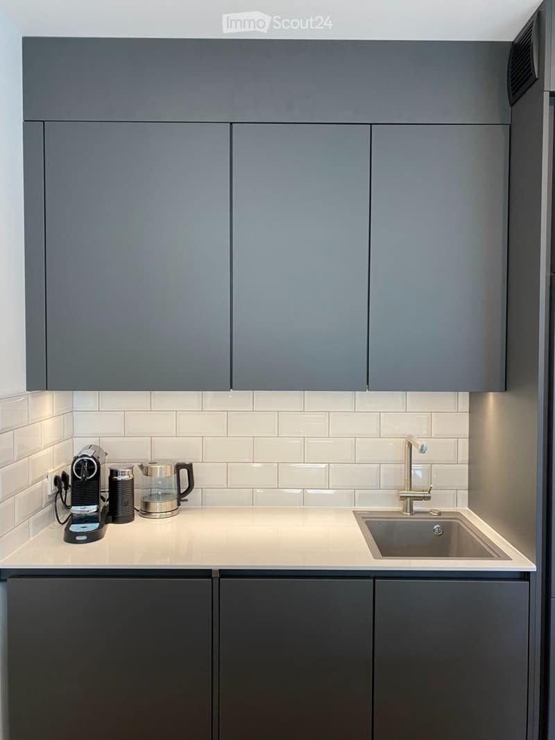 Modern kitchen with grey cabinets, white tiled backsplash, and a light-colored countertop. Features a coffee maker, electric kettle, and a modern faucet over the sink.