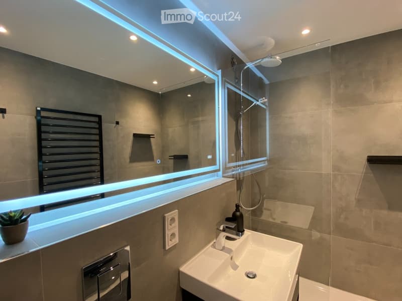 Modern bathroom with a large mirror, illuminated LED lighting, a sink with a sleek faucet, a shower area, and a radiator. The walls are tiled in a neutral tone, and a small potted plant adds a touch of greenery.