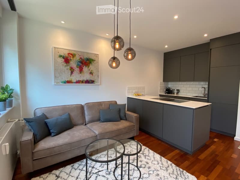 Modern open-plan living and kitchen area with grey sofa, glass coffee table, pendant lights, kitchen island, and a framed world map on the wall.
