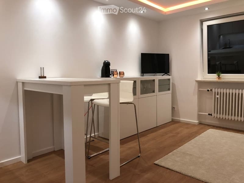 Modern, minimalist room with a white table, two white stools, a cabinet with glass doors, a television, a coffee maker, and a small potted plant on the windowsill. The room features wooden flooring and recessed lighting.