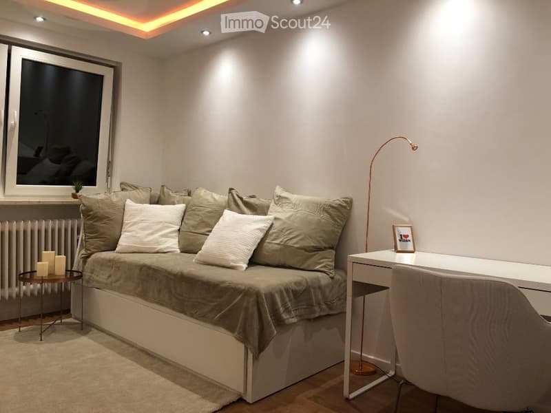 Modern, compact living space with a sofa bed, desk, chair, small table, and floor lamp. Features large window, neutral tones, and built-in storage under the sofa.