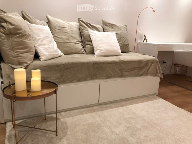 Modern bedroom with a large sofa bed, decorative pillows, a small round table with candles, a floor lamp, and a white chair.