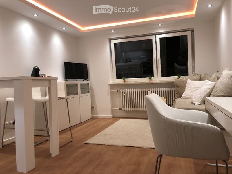 Modern living room with a sofa, coffee table, armchair, and a small dining area with a table and stools. The room features large windows, a radiator, and built-in lighting.