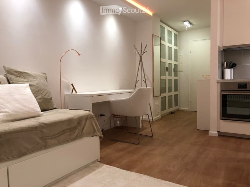 A modern, compact bedroom with a bed, desk, chair, and wardrobe. Features include a sleek white desk, a modern chair, a floor lamp, and a built-in wardrobe with glass doors. The room has light wooden flooring and white walls.