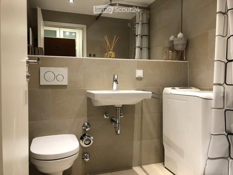Modern bathroom with beige tiles, a white sink, toilet, and shower area with a curtain. A mirror above the sink reflects decorative items on a shelf.