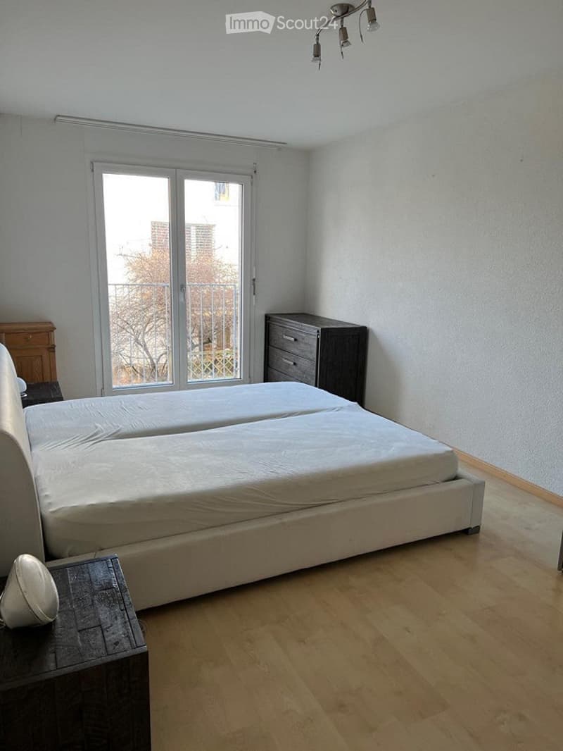 Modern bedroom with a large double bed, nightstand, drawer unit, and a chair. The room features wooden flooring and large windows with blinds.