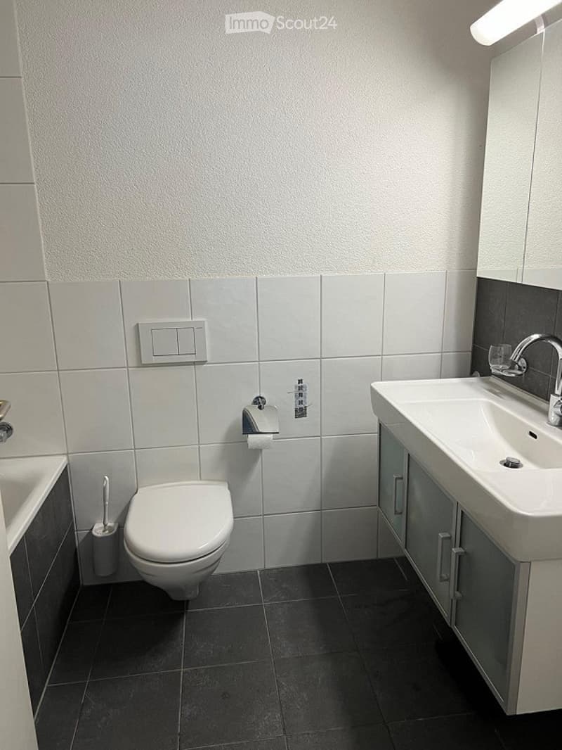 Modern bathroom with white tiled walls, dark tiled floor, toilet, sink with faucet, mirror, and light switch.