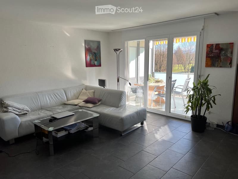 Modern living room with white leather sofa, glass coffee table, floor lamp, and large sliding glass doors leading to a balcony with outdoor furniture and scenic mountain view.