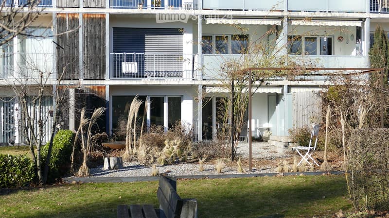 Modern two-story building with light-colored facade, large windows, and balconies. The surrounding area includes a landscaped garden with gravel pathways, tall grasses, and a few benches.