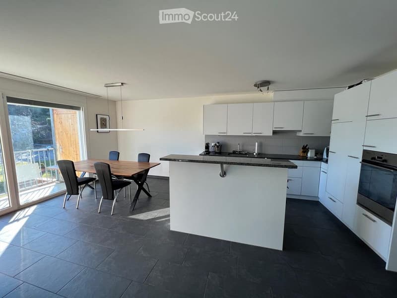 Modern kitchen with white cabinets, dark countertops, and a central island. A dining area with a wooden table and chairs is adjacent, connected to a large window with a view of the outdoor area.