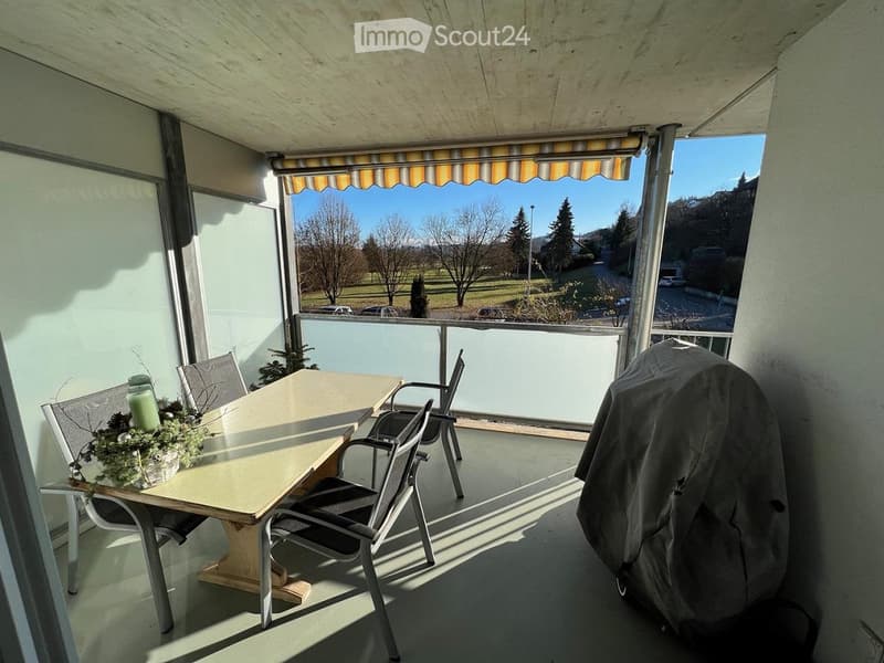 Balcony with a table, chairs, and a covered grill, overlooking a scenic landscape with trees and a clear blue sky.