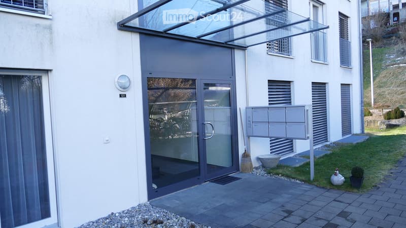Modern building entrance with glass canopy, mailboxes, small garden area with decorative items.