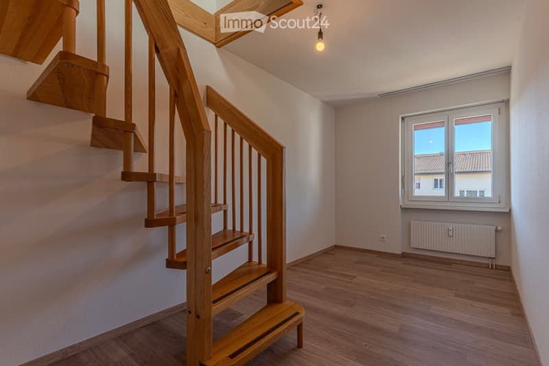 Empty room with wooden flooring, a wooden staircase leading upwards, a window with white frame, and a radiator below the window.