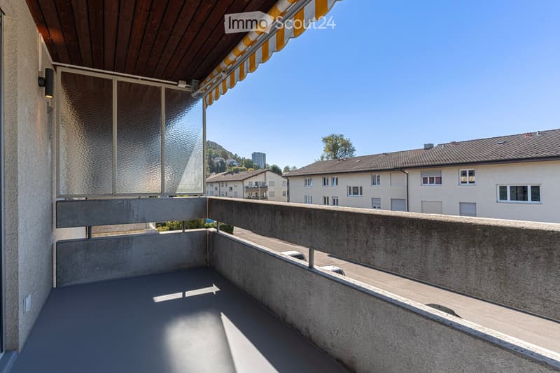 Balcony with concrete floor and railing, featuring a wooden awning and a view of neighboring buildings and greenery.