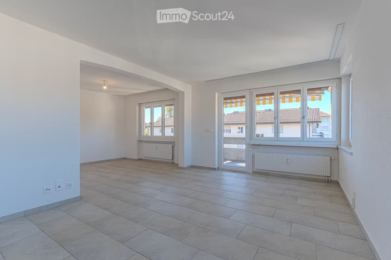 Empty room with tiled floor, white walls, large windows, radiators, and a small hallway leading to another room.
