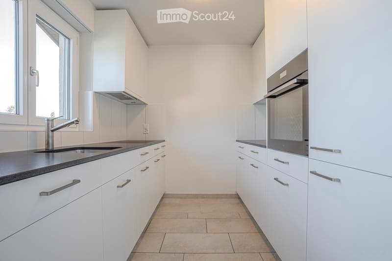 Modern kitchen with white cabinets, stainless steel sink and faucet, and built-in oven. Light-colored tiled floor and large window provide natural light.
