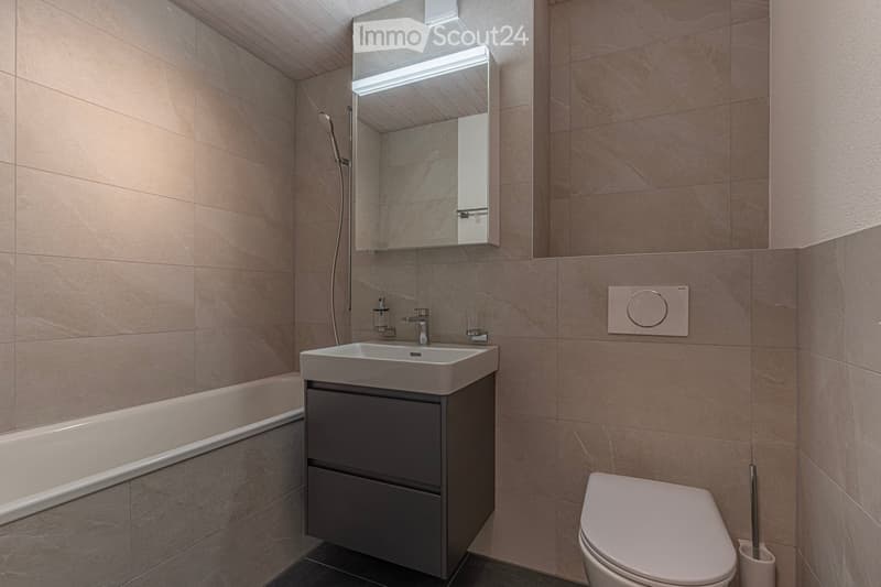 Modern bathroom with beige tiles, a white sink with a dark cabinet, a bathtub, a toilet, a mirror with built-in lighting, and a sleek faucet.