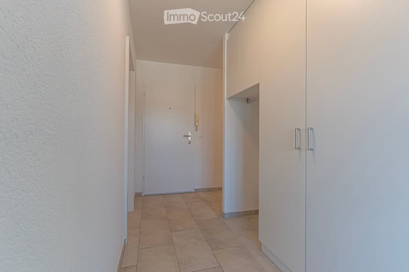 A narrow hallway with tiled flooring, white walls, and built-in white cabinets on one side. A closed white door is visible at the end of the hallway.