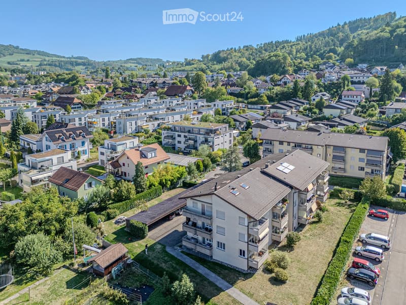 Aerial view of a residential area with multiple houses, apartments, and surrounding greenery. The buildings have varied architectural styles, with some featuring balconies and others having flat roofs.
