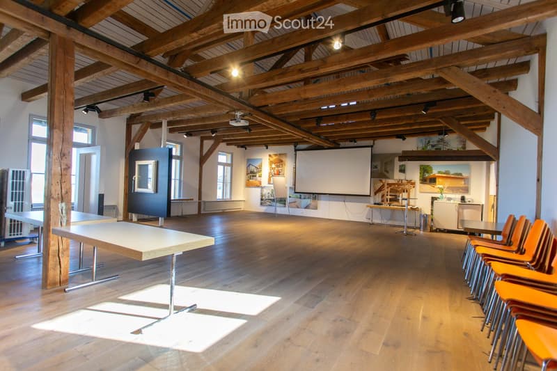 Bright room with exposed wooden beams, hardwood floor, large windows, a long table, chairs, a projector screen, and artwork on the walls.