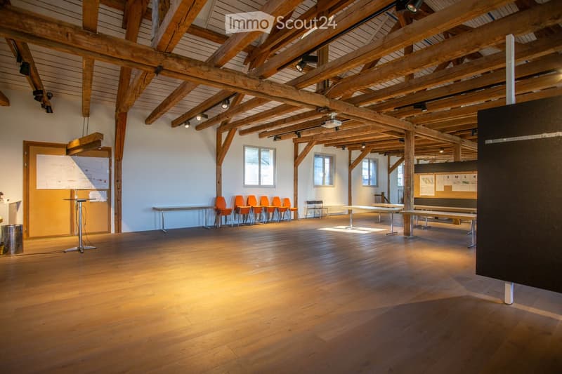 Large, open room with exposed wooden beams and polished wooden floors, featuring tables, chairs, benches, and a screen.