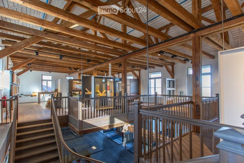 Open-plan interior with exposed wooden beams, staircase leading to an upper level, and modern furniture including a table, chairs, and a screen.