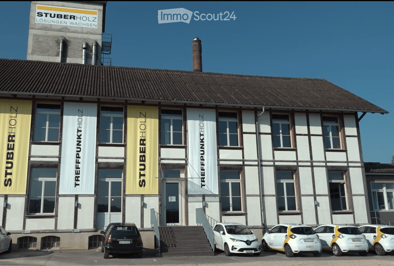 Modern industrial-style building with large windows and signage for "STUBERHOLZ" and "TREFFPUNKT HOLZ". Several cars are parked in front.