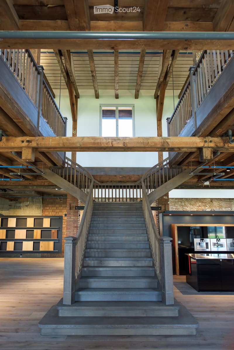 Spacious hallway with exposed wooden beams and a curved staircase leading to an upper level, featuring a modern kitchen area with stainless steel appliances visible in the background.