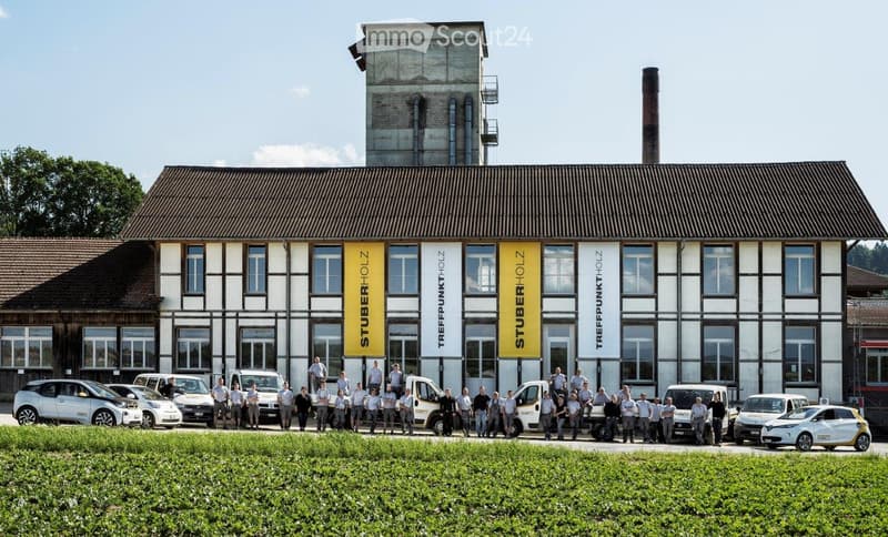 Modern industrial-style building with large windows and signage, surrounded by a group of people and parked cars.