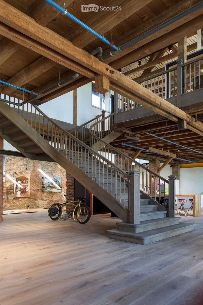 Open-concept space with exposed wooden beams, a large wooden staircase, a yellow bicycle, and two stools near a brick wall.