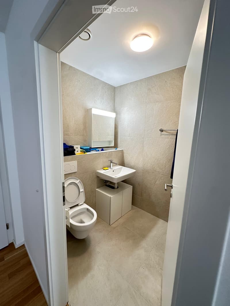 Modern, compact bathroom with beige tiles, a toilet, a sink with a countertop cabinet, and a mirror above the sink.
