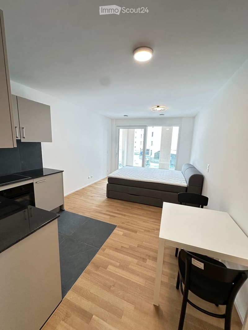 A compact studio apartment with a bed, a small table with chairs, a kitchen area with cabinets and an oven, and a large window with a view of the city.