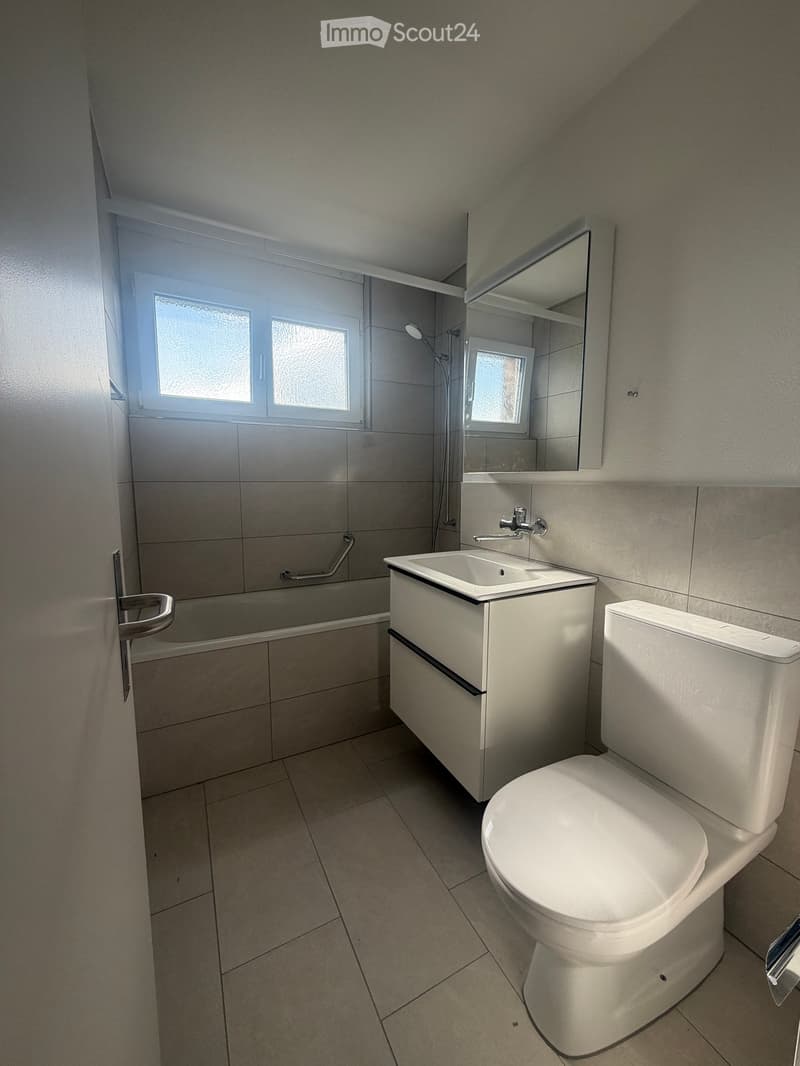 Modern bathroom with a white toilet, sink, bathtub, and mirror. The room features tiled walls and floor, with a window providing natural light.