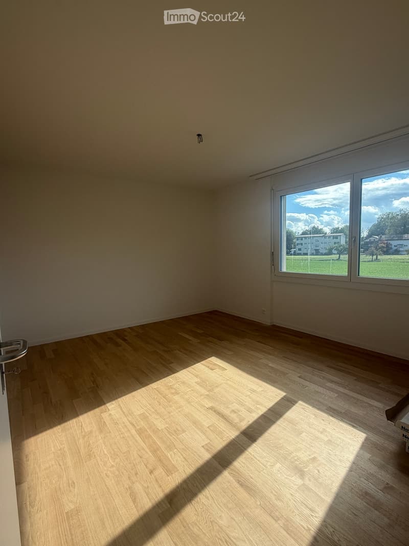 Empty room with wooden flooring and large windows letting in natural light, showing a view of a green area outside.