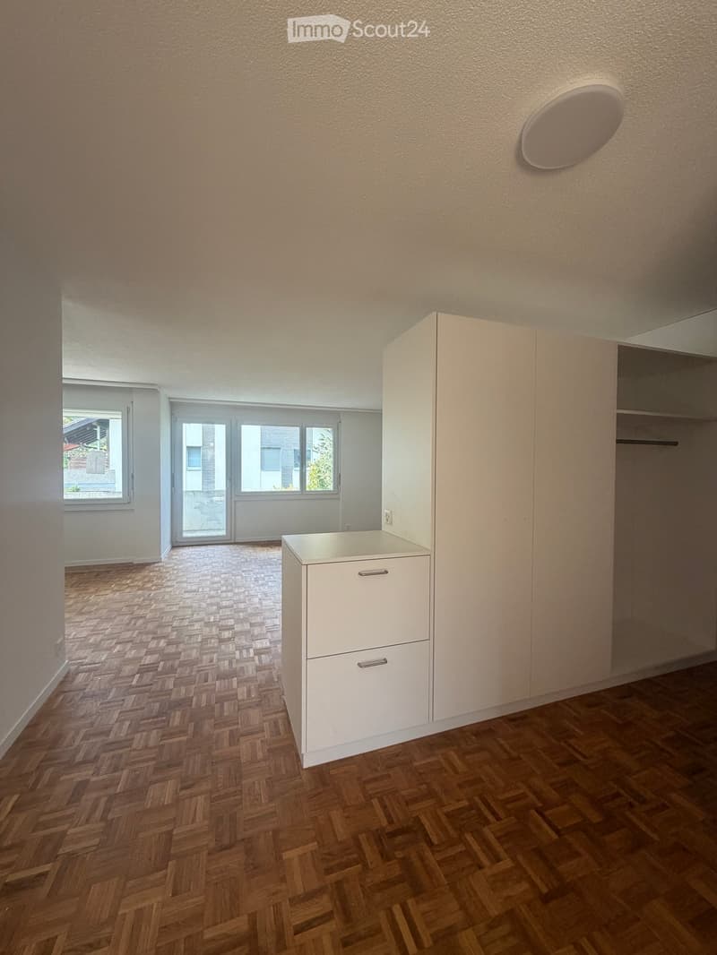 Empty room with wooden herringbone flooring, white walls, built-in closet, and a small cabinet. Large windows and sliding glass doors lead to a balcony or outdoor area.